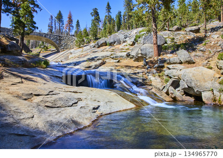 Stone Bridge Over River Cascade and Granite Rocks in Forest Landscape 134965770