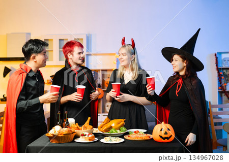 Friends in spooky Halloween costumes enjoy drinks and food at a festive indoor party with pumpkins, red cups, and laughter. 134967028