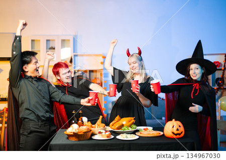 Friends in spooky Halloween costumes enjoy drinks and food at a festive indoor party with pumpkins, red cups, and laughter. Friends in spooky Halloween costumes enjoy drinks and food at a festive indoor party with pumpkins, red cups, and laughter. 134967030