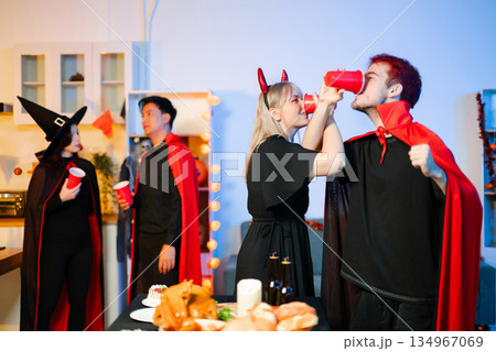 Group of young friends in Halloween costumes enjoy food and drinks at a cozy indoor party. with fun, spooky vibes, red cups, and pumpkin decor. Group of young friends in Halloween costumes enjoy food and drinks at a cozy indoor party. with fun, spooky vibes, red cups, and pumpkin decor. 134967069