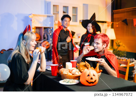 Group of young friends in Halloween costumes enjoy food and drinks at a cozy indoor party. with fun, spooky vibes, red cups, and pumpkin decor. Group of young friends in Halloween costumes enjoy food and drinks at a cozy indoor party. with fun, spooky vibes, red cups, and pumpkin decor. 134967072
