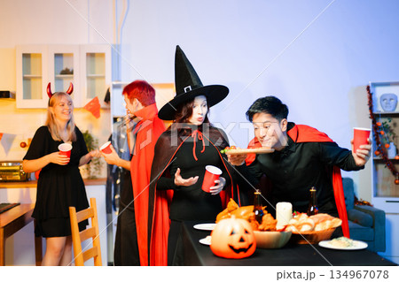 Young friends in Halloween costumes decorating pumpkin lanterns at a festive indoor party. Real people enjoying spooky vibes, red cups, snacks, and Halloween props. 134967078