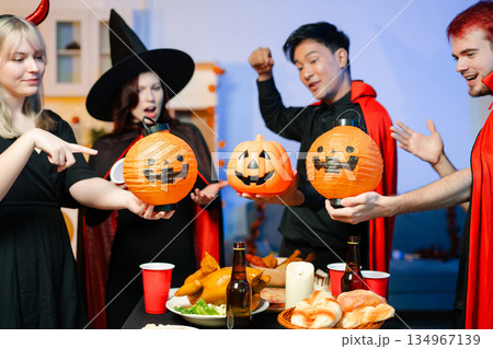 Group of friends in fun Halloween costumes laugh and play with spooky props, lanterns, and skull decor at a festive indoor party. 134967139