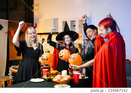Group of friends in fun Halloween costumes laugh and play with spooky props, lanterns, and skull decor at a festive indoor party. Group of friends in fun Halloween costumes laugh and play with spooky props, lanterns, and skull decor at a festive indoor party. 134967142
