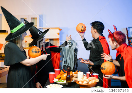Group of friends in fun Halloween costumes laugh and play with spooky props, lanterns, and skull decor at a festive indoor party. Group of friends in fun Halloween costumes laugh and play with spooky props, lanterns, and skull decor at a festive indoor party. 134967146