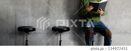 Person reading book while sitting on stool against concrete wall 134972451
