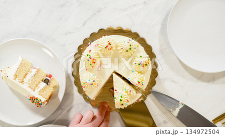 A person serves a slice of vanilla cake with sprinkles onto a white dessert plate. The cake is shown with one piece missing and sits neatly on a gold board on a marble counter. A person serves a slice of vanilla cake with sprinkles onto a white dessert plate. The cake is shown with one piece missing and sits neatly on a gold board on a marble counter. 134972504