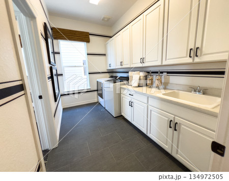 Well-lit laundry room with a large window, long countertop, sink, and white cabinetry. Decorative jars and black hardware add modern character to the space. Well-lit laundry room with a large window, long countertop, sink, and white cabinetry. Decorative jars and black hardware add modern character to the space. 134972505