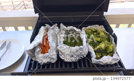 Fresh vegetables including carrots, broccoli, and asparagus are cooking in foil packets on an outdoor grill. The bright natural daylight enhances the texture and color of the vegetables in a casual 134972577