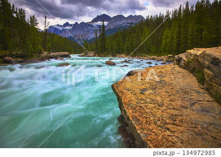 Turquoise River Water Flowing Past Rocky Ledge and Pine Forest with Mountain Peaks 134972985