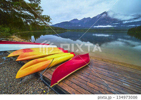 Colorful Canoes and Kayaks on Dock Reflected in Calm Lake with Majestic Mountain View Colorful Canoes and Kayaks on Dock Reflected in Calm Lake with Majestic Mountain View 134973086