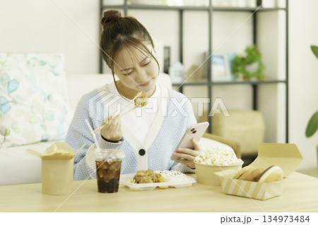 リビングでスマホを見ながらカロリーを気にしてお弁当を食べる若い日本人女性 134973484