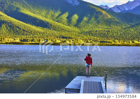 Tourist on Dock Overlooking Calm Lake and Forested Mountains in Vibrant Sunlight 134973584