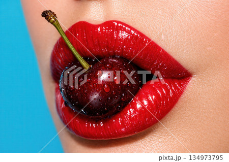 Close Up View Of Red Glossy Lips Holding A Fresh Ripe Cherry With Water Droplets On Blue Background 134973795