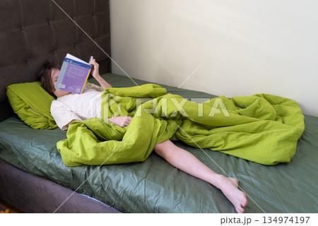 Person reading a book while lying comfortably in bed. Relaxing at home with a green duvet Person reading a book while lying comfortably in bed. Relaxing at home with a green duvet 134974197