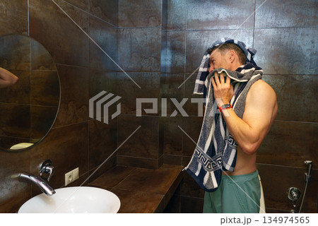 Shirtless muscular man drying his face with a towel in the bathroom. Morning hygiene and self-care Shirtless muscular man drying his face with a towel in the bathroom. Morning hygiene and self-care 134974565