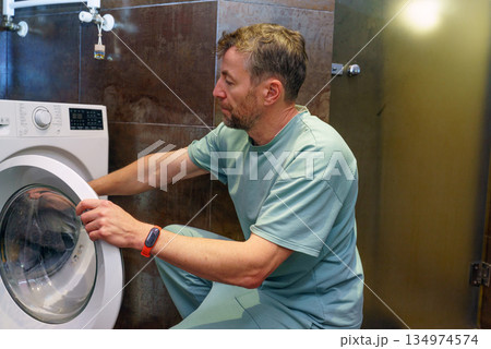 Middle-aged man loading a front-load washing machine in the bathroom. Household chores concept 134974574