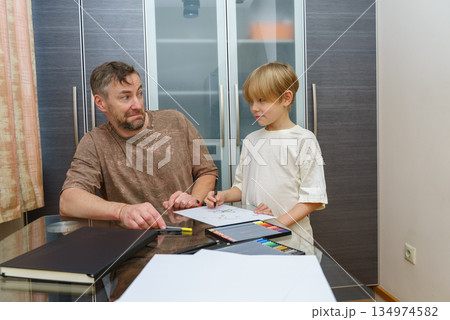 Father and son drawing together at a table. Man makes a funny face while his child creates art Father and son drawing together at a table. Man makes a funny face while his child creates art 134974582