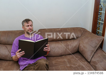A focused middle-aged man reading a book while sitting on a brown sofa. Casual lifestyle at home 134974586