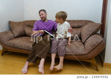 Father and son reading a book together on the couch. Parent and child bonding at home. Family quality time 134974592