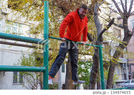 Active middle-aged man doing a muscle-up on a horizontal bar. Outdoor fitness training in autumn Active middle-aged man doing a muscle-up on a horizontal bar. Outdoor fitness training in autumn 134974655