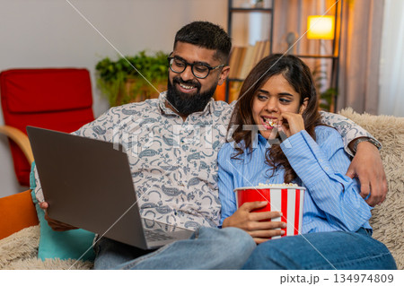 Indian family couple man woman hugging while watching series film on laptop with popcorn, enjoying 134974809