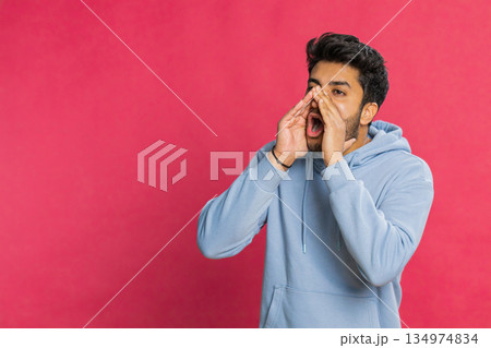 Indian young man shouting calling out loud holding hands near mouth yelling waving excited indoors Indian young man shouting calling out loud holding hands near mouth yelling waving excited indoors 134974834