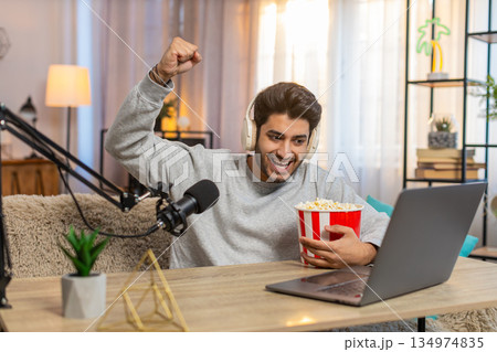 Young Indian man streaming football match commentary, eating popcorn and engaging audience at home Young Indian man streaming football match commentary, eating popcorn and engaging audience at home 134974835