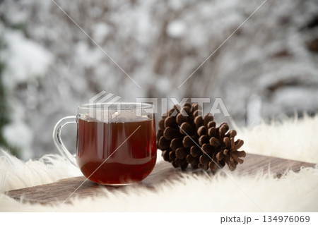 Glass cup with hot tea and steam standing on wooden board outdoors in winter next to pine cone on fluffy white fur fabric, with snowy background. Glass cup with hot tea and steam standing on wooden board outdoors in winter next to pine cone on fluffy white fur fabric, with snowy background. 134976069