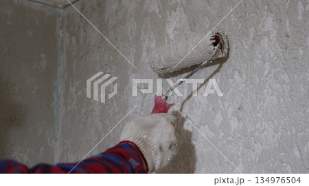 A gloved hand holds a roller with a paste solution and plasters a gray wall, close-up, covering a wall with putty using a construction roller by hand A gloved hand holds a roller with a paste solution and plasters a gray wall, close-up, covering a wall with putty using a construction roller by hand 134976504