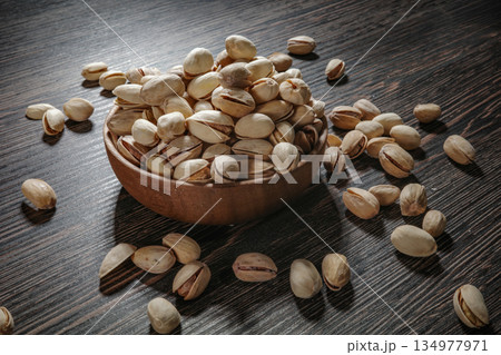 Wooden bowl filled with pistachios on dark wooden table, tasty salted nuts scattered around, healthy snack and natural food concept. 134977971