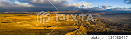Ultra high resolution panoramic view of an open pit coal mine with massive mining machines, layered terrain, and dramatic sky on the horizon. Powerful industrial landscape showing large scale Ultra high resolution panoramic view of an open pit coal mine with massive mining machines, layered terrain, and dramatic sky on the horizon. Powerful industrial landscape showing large scale 134980457