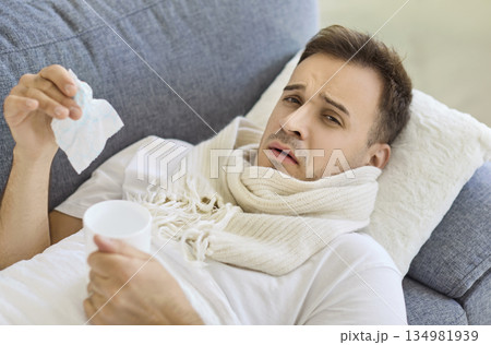 Sick man with scarf holding tissue and cup, resting on sofa at home interior Sick man with scarf holding tissue and cup, resting on sofa at home interior 134981939