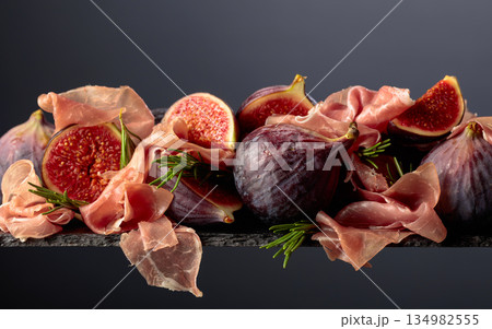 Prosciutto with rosemary and fig fruit on a black background. Prosciutto with rosemary and fig fruit on a black background. 134982555