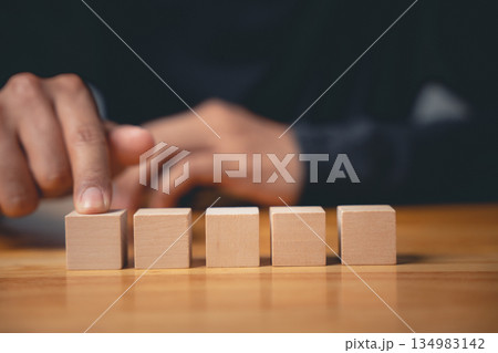 Close-up of a hand pointing at a row of five wooden blocks on a wooden table. 134983142