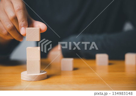 A hand placing a wooden block on top of a stack of two, on a wooden table. More blocks are out of focus in the background. A hand placing a wooden block on top of a stack of two, on a wooden table. More blocks are out of focus in the background. 134983148
