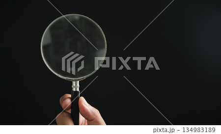 A hand holding a magnifying glass against a black background. The magnifying glass is circular and has a black handle. A hand holding a magnifying glass against a black background. The magnifying glass is circular and has a black handle. 134983189