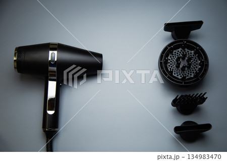 A black hair dryer on one side, on the other there are different drying attachments - a comb, a diffuser and classic nozzles of different diameters on a gray background, top view. The concept of 134983470