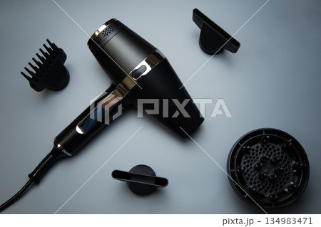 A black hair dryer around which there are different drying attachments - a comb, a diffuser and classic nozzles of different diameters on a gray background, top view. High quality photo A black hair dryer around which there are different drying attachments - a comb, a diffuser and classic nozzles of different diameters on a gray background, top view. High quality photo 134983471