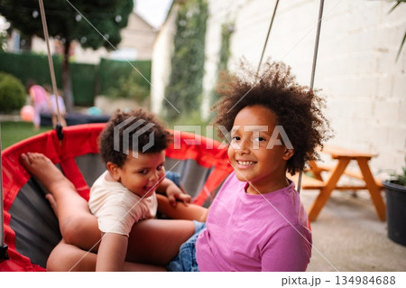 Siblings sharing swing in backyard enjoying childhood playtime Siblings sharing swing in backyard enjoying childhood playtime 134984688