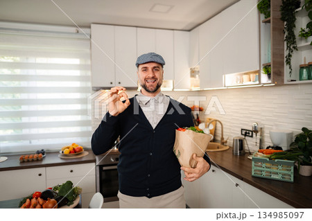 Happy man holding shopping bag with groceries in kitchen Happy man holding shopping bag with groceries in kitchen 134985097
