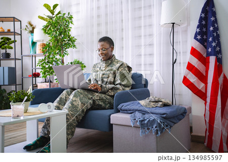 Female soldier sitting on sofa using laptop with United States flag in background 134985597