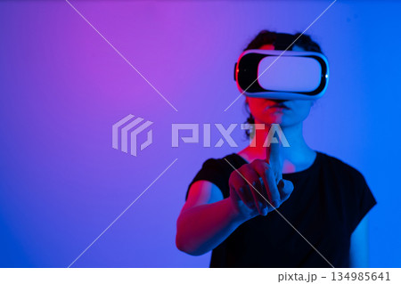 Close up of woman wearing virtual reality headset and touching virtual interface with finger in futuristic neon lighting 134985641