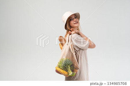 Young woman standing in profile with reusable mesh shopping bag full of vegetables and bread, sustainable food shopping and eco conscious lifestyle concept 134985708