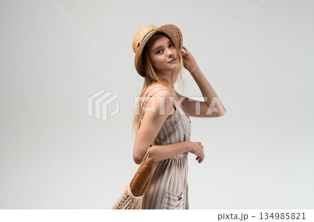 Young woman in straw hat adjusting hair while holding reusable mesh shopping bag with bread, eco friendly lifestyle portrait, sustainable grocery shopping 134985821