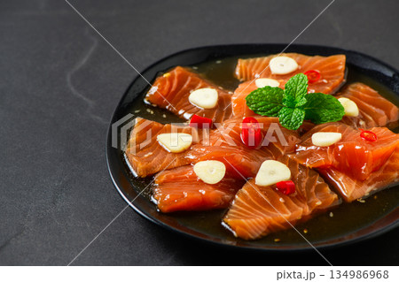 Korean-style soy marinated salmon slices served with garlic, red chili, and herbs on a black plate. Popular Korean food known as salmon jang, premium seafood dish Korean-style soy marinated salmon slices served with garlic, red chili, and herbs on a black plate. Popular Korean food known as salmon jang, premium seafood dish 134986968