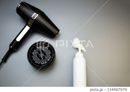 A top view of hair styling foam in a white bottle, next to it is a hairdryer with a diffuser nozzle on a gray background with space for text. The concept of long-term hair styling, creating hairstyles A top view of hair styling foam in a white bottle, next to it is a hairdryer with a diffuser nozzle on a gray background with space for text. The concept of long-term hair styling, creating hairstyles 134987079