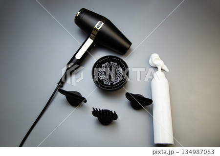 A black hair dryer with different attachments - a diffuser for creating curls and curls, a comb and classic lie on a gray background next to a hair mousse for long-lasting styling with white foam on A black hair dryer with different attachments - a diffuser for creating curls and curls, a comb and classic lie on a gray background next to a hair mousse for long-lasting styling with white foam on 134987083