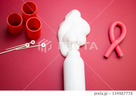Close-up view from above in the center of a bottle of foam for long-lasting hair styling lies on a pink background, white foam poured out of it, round curlers and a curved pink curler lie nearby. The 134987778