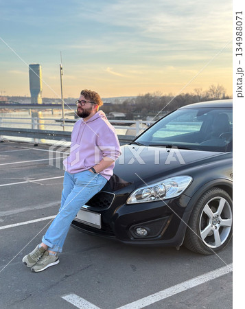 Man leaning on car near river at sunset. Urban lifestyle, freedom, modern masculinity, and reflective pause in city environment 134988071